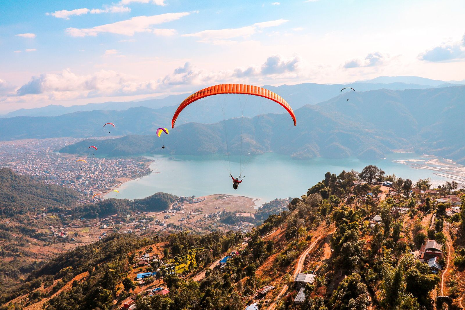 A paraglider with an orange wing flying above Phewa Lake and Pokhara